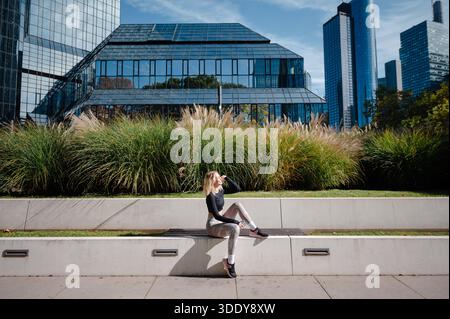 Una donna bionda in abbigliamento fitness si prende una pausa su una panchina di cemento, circondata da lussureggianti erbe ornamentali e moderni edifici in vetro Foto Stock