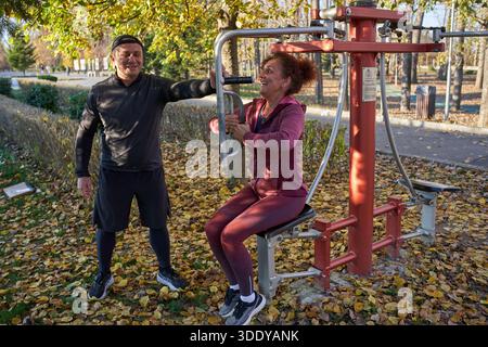 L'uomo aiuta la donna a utilizzare le attrezzature fitness all'aperto nel parco autunnale coperto di foglie gialle cadute Foto Stock