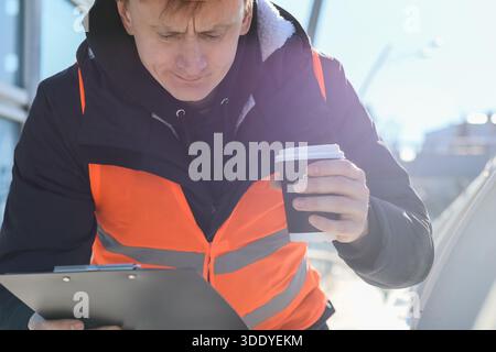 Un giovane maschio con elmetto e giubbotto tiene la bevanda mentre studia documenti sulle infrastrutture urbane innevate Foto Stock