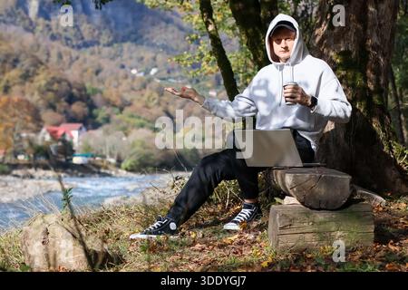 Un giovane maschio sorridente con felpa bianca si muove apertamente tenendo la tazza accanto al laptop su un tronco di legno in un ambiente montano Foto Stock