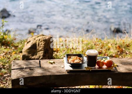 Piatto a grana in contenitore nero con forchetta in legno, tazza da caffè scuro con coperchio bianco, due frutti arancioni, foglia gialla su tavoletta bianca su banco in legno con la Foto Stock