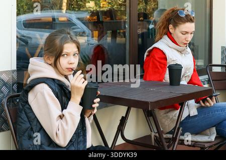 Donna adulta e preadolescente siedono insieme al caffè all'aperto, entrambi con tazze nere, donna che controlla il telefono in un ambiente informale. Foto Stock