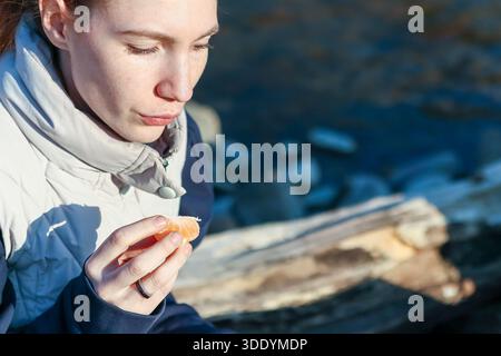 Giovane donna in mantello beige che profuma di arancio mandarino aromatico all'aperto, vicino al fiume calmo in autunno Foto Stock