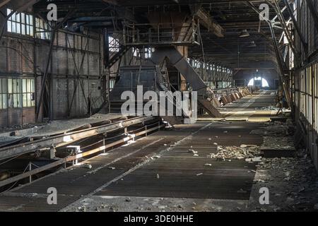 Ampia visuale di un locale industriale abbandonato con grandi strutture metalliche per l'installazione del trasportatore e detriti sul pavimento Foto Stock
