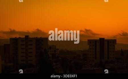 Foto orizzontale grandangolare di un cielo arancione luminoso al tramonto con sagome scure di edifici residenziali e nuvole che coprono il sole Foto Stock