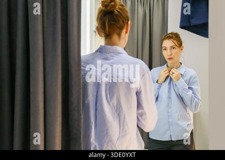 La persona in camicetta a righe esamina l'abbigliamento a specchio della cabina di montaggio con tende e grucce Foto Stock