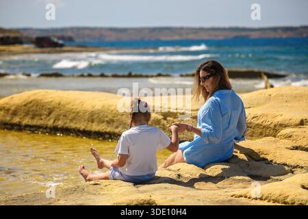 Una donna e una ragazza si siedono sulle rocce calcaree gialle in riva al mare, tenendosi per mano e spruzzando acqua con i piedi sotto il sole luminoso, vista sul retro. Foto Stock