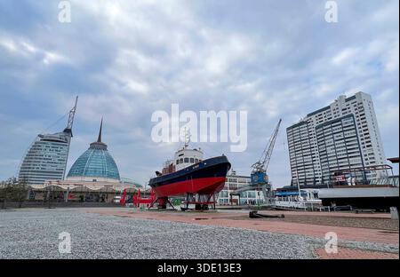 Bremerhaven, Deutschland, Europa. Außenansicht des Museumshafens mit dem historischen Schiff Seute Deern aufgebockt an Land, umgeben von Hafeninfrastruktur. Im Hintergrund moderne Wahrzeichen wie das KlimaHaus Bremerhaven und das Atlantic Hotel Sail City, Hafenkräne sowie Hochhausbebauung. Weitwinkelaufnahme aus niedriger Perspektive, offener Platz mit Pflaster und Schotter, bewölkter Himmel. Stadtentwicklung, Tourismus, Maritime Geschichte, Architektur, Gesellschaft, Hafen, Symbolfoto. *** Bremerhaven, Germania, Europa Vista esterna del porto del museo con la storica nave Seute Deern jacke Foto Stock