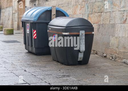 Modern Street Waste Containers contro Old Wall; due grandi contenitori di plastica nera e blu posizionati su una strada bagnata e acciottolata accanto a una A. Foto Stock