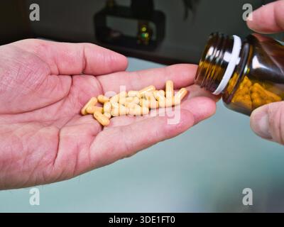 Versare in mano le capsule gialle dal flacone per pillole; primo piano di una persona che versa capsule gialle/abbronzate da un flacone di vetro ambrato nel Foto Stock