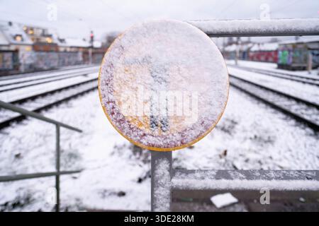 Primo piano di un cartello di avvertimento coperto di neve alla stazione di Colonia Ehrenfeld. Binari ferroviari sfocati e atmosfera invernale sullo sfondo Foto Stock