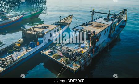 Balikpapan, Indonesia - 8 novembre 2025. un pescatore in piedi sul ponte disordinato della sua tradizionale barca da pesca in legno con un pannello solare sul Foto Stock