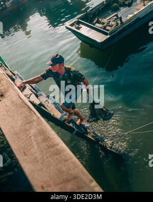 Balikpapan, Indonesia - 8 novembre 2025. Un pescatore a piedi nudi, in camicia camo, sale dal mare su un molo una scala di legno improvvisata. Porta con sé Foto Stock