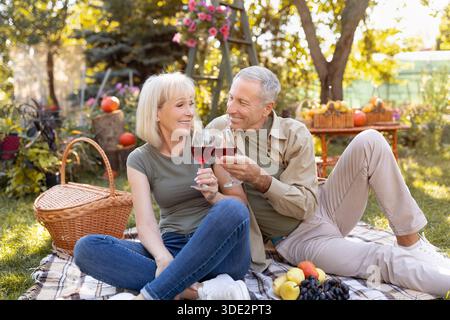 Festeggiamo l'anniversario. Amorevole coppia anziana bere vino, seduta su coperta mentre si ha pic-nic in giardino Foto Stock