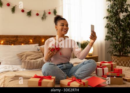 Festa di Natale online. Eccitata signora nera usando il cellulare, prendendo selfie o facendo videochiamata, celebrando le feste di Natale a casa Foto Stock