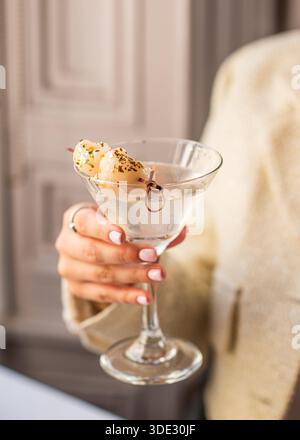 Le mani di una donna tengono in mano un martini di litchi servito in un classico bicchiere di martini. La bevanda chiara e dai colori chiari trasmette eleganza e freschezza. Foto Stock