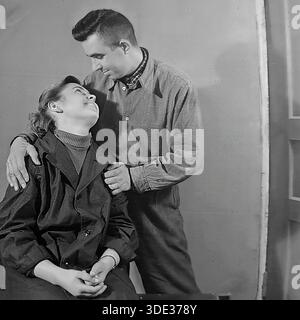 Slaviansk, regione di Donetsk, Ucraina, URSS, anni 1950 Un coinvolgente ritratto d'archivio vintage in bianco e nero che cattura un tenero momento romantico tra una giovane coppia sovietica, Mikhail Ilyich Kravelis e sua moglie Valentina Timofeevna Kravelis. Il marito sta in piedi e abbraccia delicatamente la moglie seduta, guardandola negli occhi con profondo affetto e un sorriso sincero. Sono vestiti con un tipico modesto abbigliamento casual del dopoguerra, con Mikhail in una camicia con bottoni scuri e Valentina in un cappotto su un maglione a collo alto. L'immagine irradia felicità, giovinezza e amore autentico all'interno di una famiglia Foto Stock