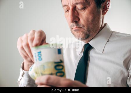 Uomo d'affari in camicia bianca contando con ansia un rotolo spesso di 100 euro di banconote con sopracciglia a solco. Messa a fuoco selettiva. Foto Stock
