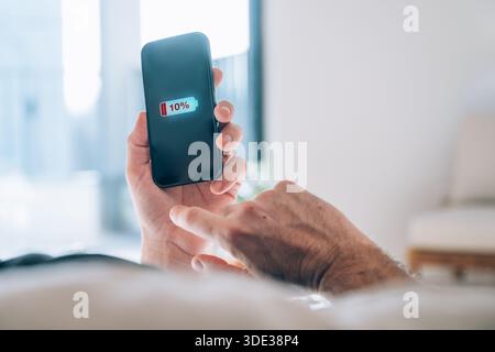 Primo piano di uno smartphone nero con mano maschio che mostra un avviso critico della batteria del 10%. Bassa ansia della batteria, dipendenza da dispositivi mobili, stress tecnologico, urgenza Foto Stock