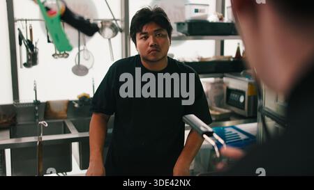 Un giovane chef asiatico stressato che cucina in una cucina buia preparando cibo da ristorante Foto Stock