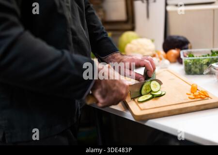Primo piano di uno chef professionista che prepara gli ingredienti per i pasti vegetariani nella cucina del ristorante. Sta tagliando il cetriolo. Foto Stock