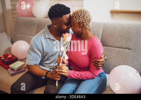 Felice coppia amorevole che festeggia il compleanno della donna a casa. Il fidanzato sta dando la rosa alla sua ragazza e la sta abbracciando. Foto Stock