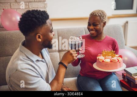 Felice giovane coppia che festeggia il compleanno della donna a casa. La donna tiene la torta e brindano con il vino. Foto Stock
