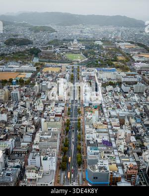 Vista aerea di una lunga strada che conduce al castello di Himeji circondato da un fitto paesaggio urbano, con montagne sullo sfondo, Himeji, Hyogo, Giappone. Foto Stock
