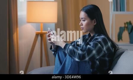 Vista laterale donna asiatica triste seduta sul divano di casa con smartphone infelice ragazza coreana cinese che scorre sfogliando l'app del telefono cellulare guardare Foto Stock
