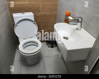 Interno del bagno con WC in ceramica bianca, coperchio aperto e lavandino a parete. Rubinetto cromato, sapone, deodorante per ambienti. Spazzolina per WC, contenitore della spazzatura. Muro Foto Stock