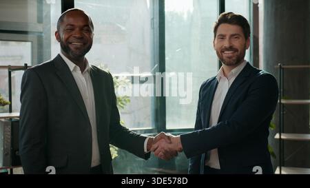 Due uomini d'affari multinazionali team uomini d'affari che stringono la mano guardando la macchina fotografica sorridente allegro lavoro di squadra Caucasico candidato maschio con Foto Stock