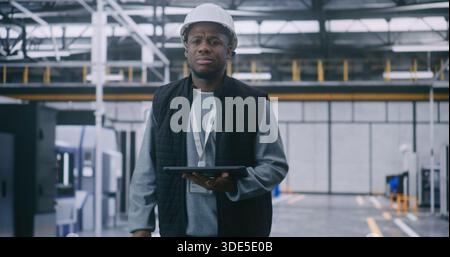 Ingegnere con Hard Hat che controlla i dati sul tablet. L'ingegnere maschio fornisce l'ispezione di fabbrica. Un uomo entra in una grande e moderna fabbrica industriale. Concept Mobile Management, sicurezza e supervisione operativa. Foto Stock