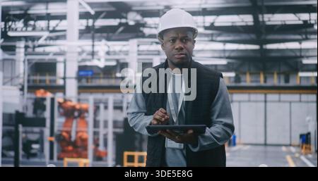 Ingegnere con Hard Hat che controlla i dati sul tablet. L'ingegnere maschio fornisce l'ispezione di fabbrica. Un uomo entra in una grande e moderna fabbrica industriale. Concept Mobile Management, sicurezza e supervisione operativa. Foto Stock