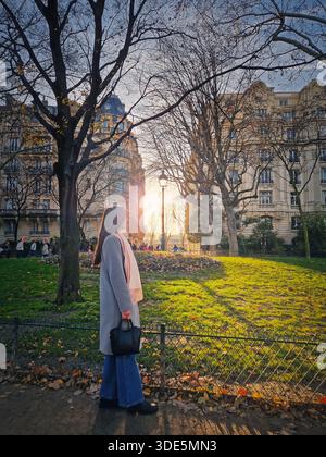 Bella giovane donna che cammina attraverso un parco parigino illuminato dal sole, vestita con un cappotto grigio mentre passeggia oltre le foglie autunnali cadute verso il sole splendente Foto Stock