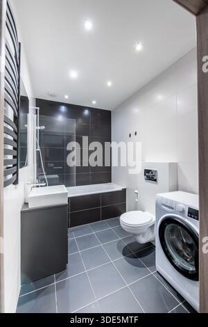 Bagno moderno e luminoso con elegante arredo bianco e nero, vasca, WC e lavatrice Foto Stock