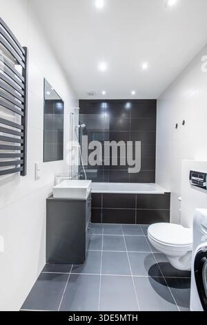 Bagno moderno e luminoso con elegante arredo bianco e nero, vasca, WC e lavatrice Foto Stock