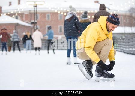 Uomo di mezza età caucasico che lega pattini neri sulla pista di pattinaggio su ghiaccio in giornate invernali innevate all'aperto Foto Stock