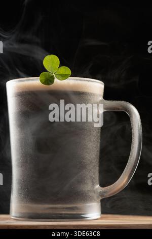 Tazza di birra scura e robusta con foglia di trifoglio di roccia e misterioso fumo su sfondo nero per il giorno di San Patrizio Foto Stock