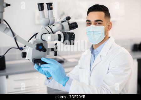 Dentista maschio arabo che indossa maschera medica protettiva usando microscopio dentale in clinica Foto Stock