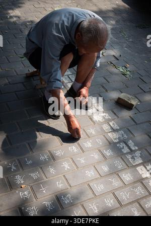 Uomo anziano che pratica la tradizionale calligrafia acquatica su un marciapiede, scrivendo caratteri cinesi usando acqua e un pennello. Street art Pechino Cina Foto Stock
