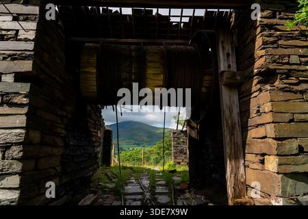 Interno di un edificio abbandonato in una cava di ardesia con un grande tamburo avvolgente e cavo in acciaio, che incornicia la montagna lontana attraverso la porta Foto Stock