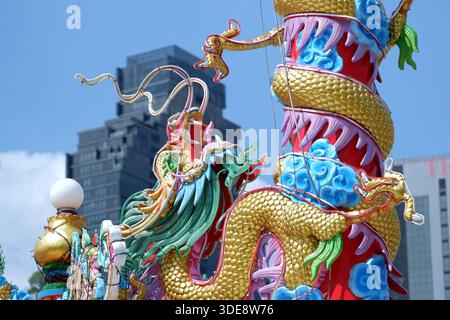 Una vista ravvicinata mostra una testa di drago su un palo cerimoniale contro il cielo azzurro del cimitero di Teochew. Sullo sfondo appare un moderno grattacielo di Bangkok. Foto Stock
