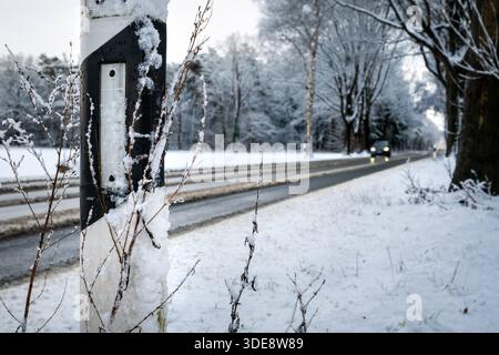 Winterlicher Straßenverkehr auf Landstraße in Niedersachsen Hude/Oldenburg, Deutschland, 03.01.2026: Ein beschneiter Leitpfosten steht am Rand einer winterlichen Landstraße. Die Fahrbahn ist teilweise von Schnee und Schneematsch bedeckt, im Hintergrund nähert sich ein Fahrzeug mit eingeschaltetem Licht. Bäume entlang der Straße sind von Frost überzogen. Symbolbild. *** Traffico stradale invernale su una strada di campagna nella bassa Sassonia Hude Oldenburg, Germania, 03 01 2026 Un delineatore innevato si trova al bordo di una strada di campagna ventosa la strada è parzialmente coperta di neve e ghiaia, sullo sfondo a Foto Stock