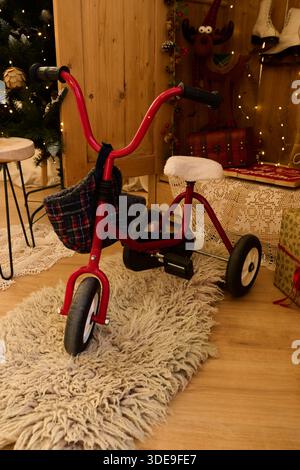 Un triciclo rosso vintage con sedile a plaid e cestino, posto su un tappeto accogliente in una sala festiva decorata con luci e ornamenti natalizi Foto Stock