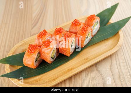 I panini di sushi fatti con salmone e uova sono posti su un tavolo di legno con foglie verdi. Il piatto mette in risalto la preparazione e la presentazione del sushi. Foto Stock