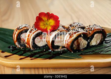 Gli involtini di sushi sono disposti ordinatamente su un piatto di legno. I rotoli presentano vari ripieni e sono ricoperti da semi di sesamo e un fiore luminoso. Un gocciolamento Foto Stock