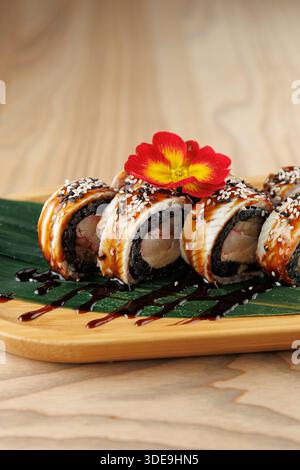 Un piatto di legno mostra involtini di sushi meticolosamente preparati con salsa lucida, semi di sesamo spolverati e una vivace decorazione floreale. Questo piatto Highli Foto Stock