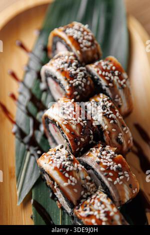 Sushi Rolls con anguilla e sesamo seduti su un piatto di bambù. La presentazione include foglie verdi e un filo di salsa per un'esaltazione del gusto. Foto Stock