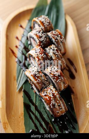 Un piatto di legno mostra sushi panini pieni di anguilla e guarniti con semi di sesamo. Gli involtini si adagiano su foglie di banana con un filo di salsa, creatina Foto Stock