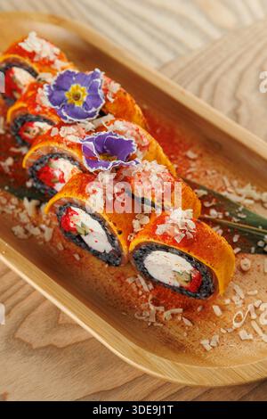 Un piatto di legno mostra colorati sushi roll ripieni di formaggio spalmabile e verdure, ricoperti di fiori e decorazioni. Le caratteristiche principali della presentazione Foto Stock